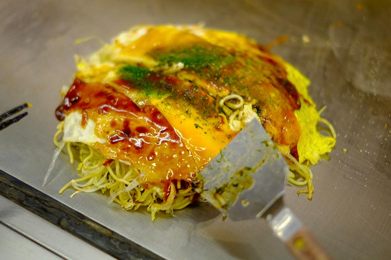 Okonomiyaki, Hiroshima-style, includes a thin crêpe-like outer layer, tons of cabbage, noodles, various seafoods, and numerous sauces. Okonomiyaki, Hiroshima-style, includes a thin crêpe-like outer layer, tons of cabbage, noodles, various seafoods, and numerous sauces.